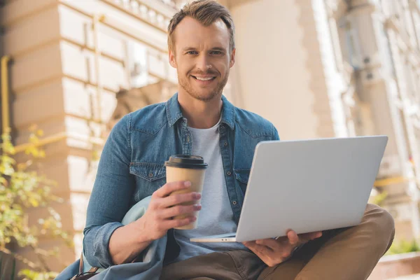 Happy young man with paper cup of coffee using laptop on street and looking at camera — Stock Photo