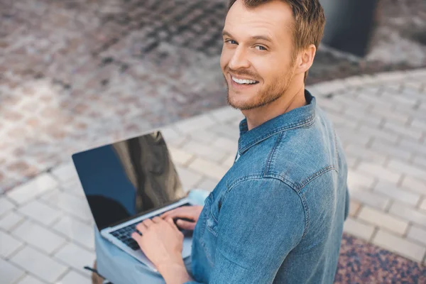 High angle view of handsome young man using laptop with blank screen on street and looking at camera — Stock Photo