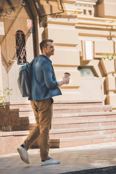 Jovem casual com xícara de papel de café e mochila andando pela rua e olhando para longe — Fotografia de Stock