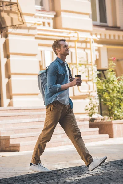 Casual young man with coffee to go and backpack walking by street and looking away — Stock Photo