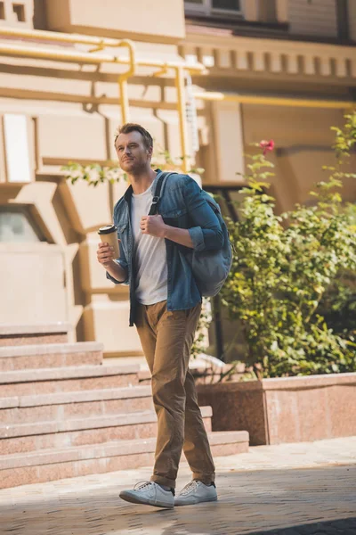 Jovem bonito com café para ir e mochila andando pela rua e olhando para longe — Fotografia de Stock