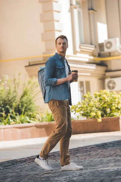 Jovem bonito com café para ir e mochila andando pela cidade e olhando para longe — Fotografia de Stock