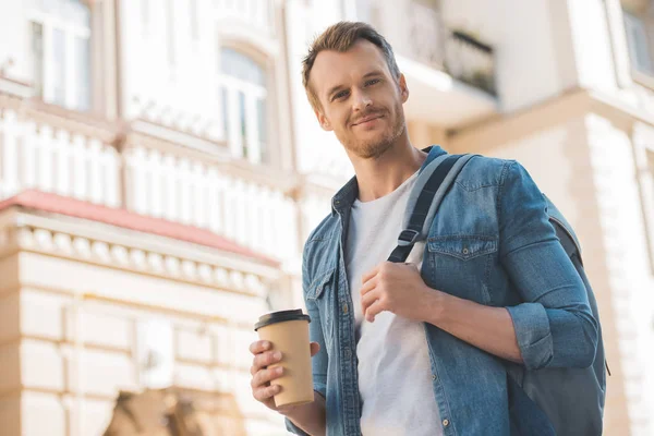 Bottom view of handsome young man with coffee to go and backpack walking on street and looking at camera — Stock Photo
