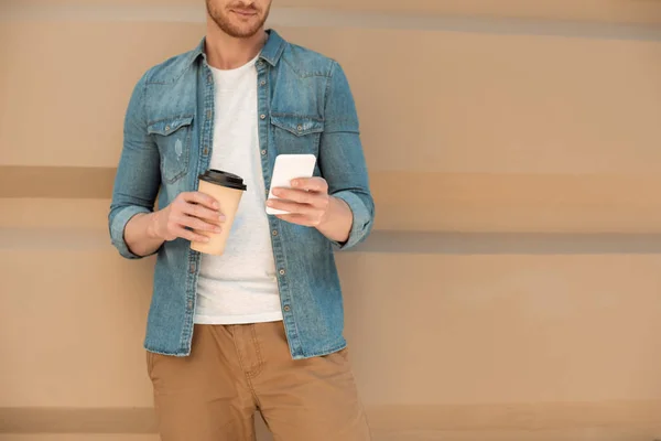 Обрезанный снимок молодого человека с кофе идти с помощью смартфона перед стеной — стоковое фото