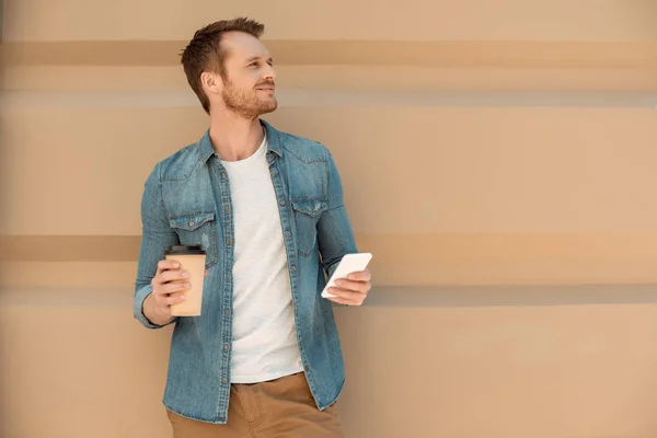 Sorrindo jovem com café para ir usando smartphone na frente da parede — Fotografia de Stock