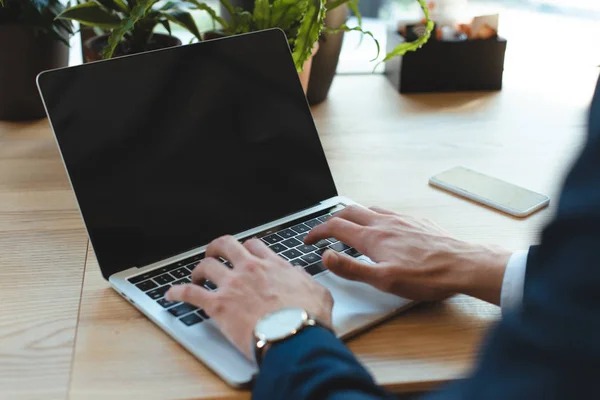 Plan recadré de l'homme d'affaires tapant sur ordinateur portable avec écran blanc à la table avec smartphone dans le café — Photo de stock