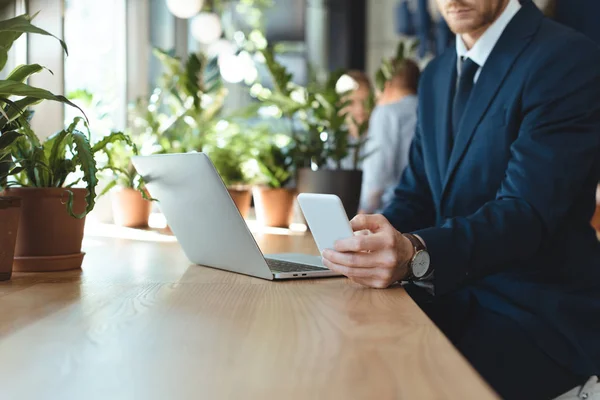 Partial view of businessman with smartphone sitting at table with laptop in cafe — Stock Photo