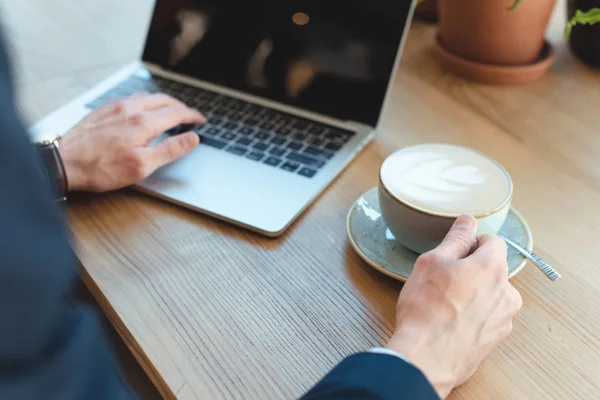 Cropped shot of businessman with cup of coffee working on laptop with blank screen in cafe — Stock Photo