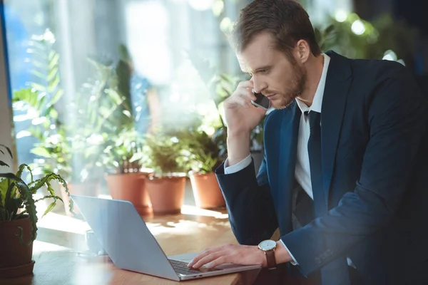 Vista lateral de hombre de negocios con estilo en traje hablando en el teléfono inteligente mientras se trabaja en el ordenador portátil en la cafetería - foto de stock