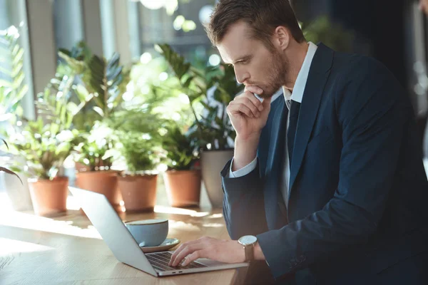 Side view of pensive businessman working on laptop at table with cup of coffee in restaurant — Stock Photo