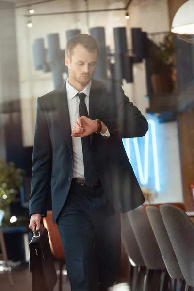 Businessman in fashionable suit with suitcase checking time while walking in cafe — Stock Photo