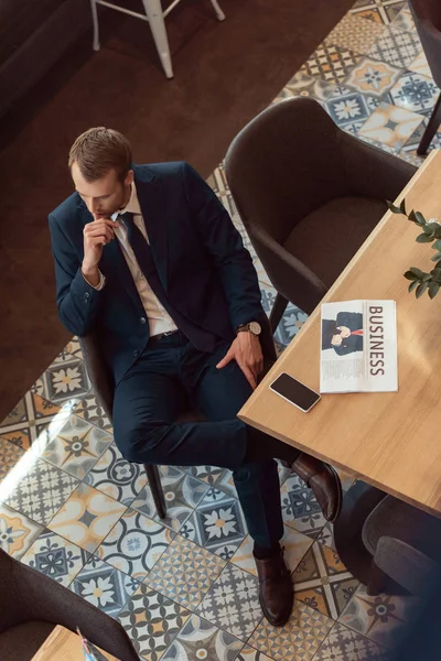 Vista aérea del hombre de negocios en traje con periódico en la mesa con taza de café y teléfono inteligente en la cafetería - foto de stock