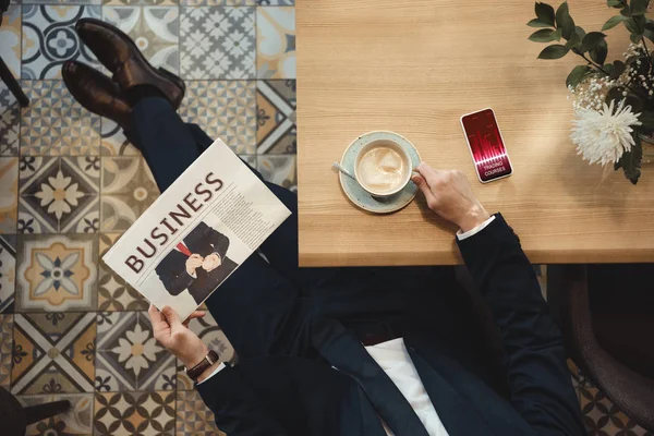 Overhead view of businessman with newspaper sitting at table with cup of coffee and smartphone in cafe — Stock Photo