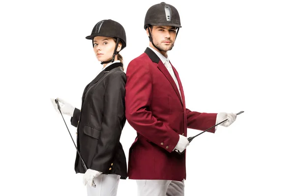 Serious young equestrians in uniform and helmets holding horseman sticks while leaning back to back and looking at camera isolated on white — Stock Photo