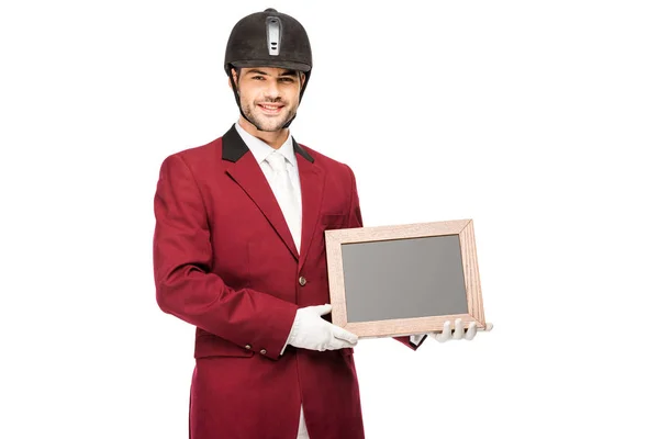 Happy young horseman in uniform holding blank chalkboard and looking at camera isolated on white — Stock Photo