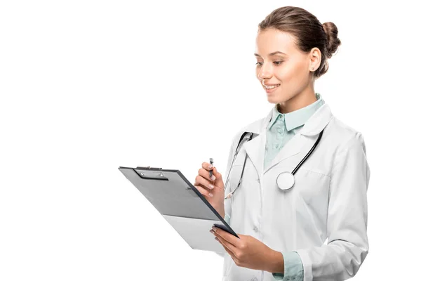 Happy young female doctor in medical coat writing in clipboard isolated on white — Stock Photo
