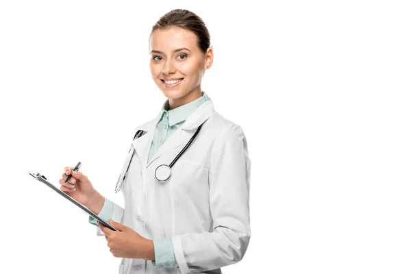 Sonriente joven doctora en abrigo médico escrito en portapapeles aislado en blanco — Stock Photo