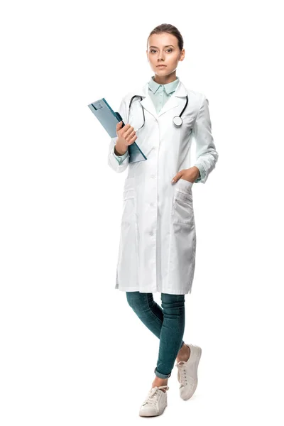 Confident young female doctor in medical coat holding clipboard isolated on white — Stock Photo