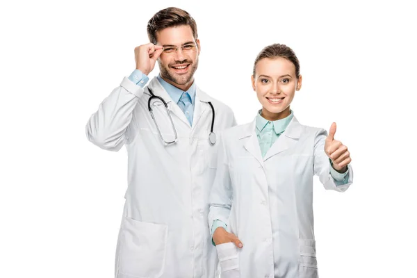 Atractiva doctora haciendo gesto pulgar hacia arriba mientras su colega masculino de pie cerca aislado en blanco - foto de stock