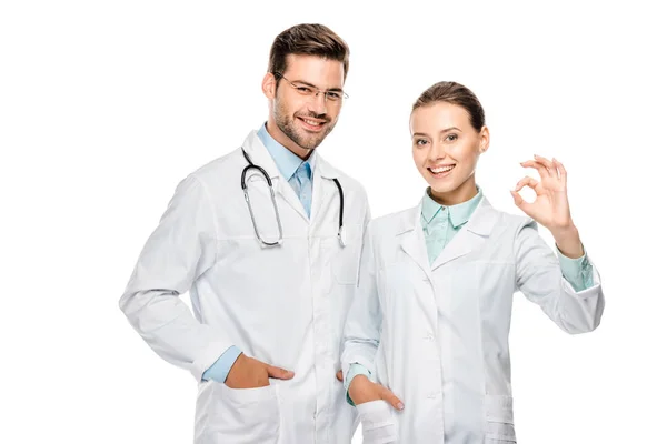 Hermosa mujer médico mostrando ok signo mientras que su colega masculino de pie cerca con estetoscopio aislado en blanco — Stock Photo