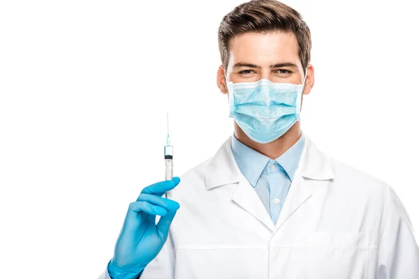 Alegre joven médico masculino en guantes de látex y máscara médica que sostiene la jeringa aislada en blanco - foto de stock