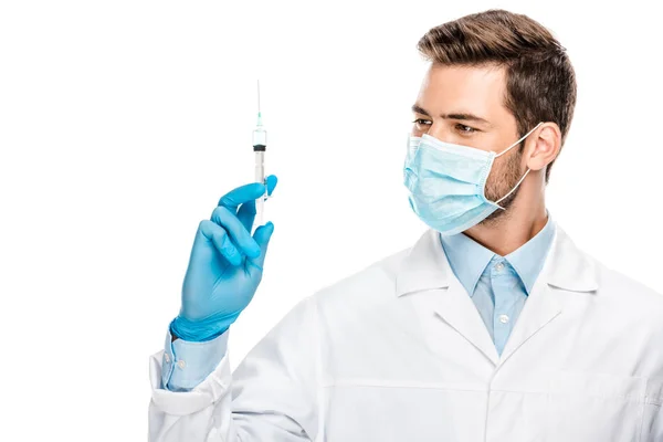 Sonriente joven doctor en guantes de látex y mascarilla médica sosteniendo jeringa aislada en blanco - foto de stock