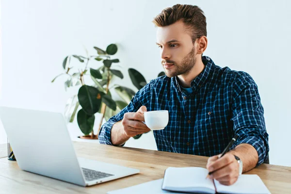 Jovem empresário com xícara de café participando de webinar na mesa com laptop no escritório — Fotografia de Stock