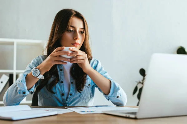 Портрет концентрованої бізнес-леді з чашкою кави на робочому місці з паперами та ноутбуком в офісі — Stock Photo