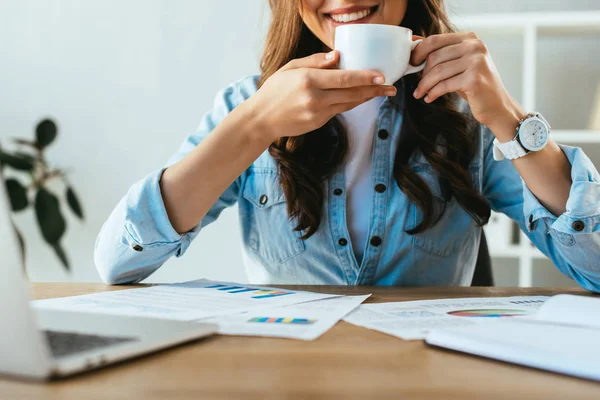 Colpo ritagliato di donna d'affari sorridente con tazza di caffè sul posto di lavoro con documenti e laptop in ufficio — Foto stock