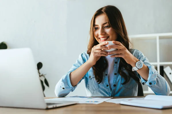 Retrato de mujer de negocios sonriente con taza de café en el lugar de trabajo con papeles y computadora portátil en la oficina — Stock Photo