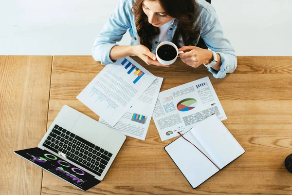 Вид сверху на деловую женщину с чашкой кофе на рабочем месте с бумагами и ноутбуком в офисе — стоковое фото