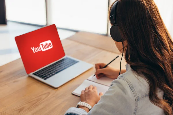 Partial view of businesswoman in headphones sitting at workplace with notebook and laptop with youtube website — Stock Photo