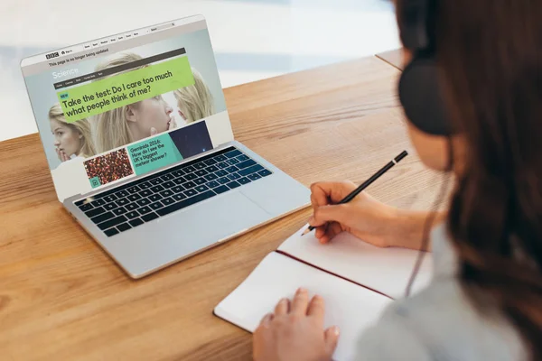 Partial view of businesswoman in headphones sitting at workplace with notebook and laptop with bbc website — Stock Photo