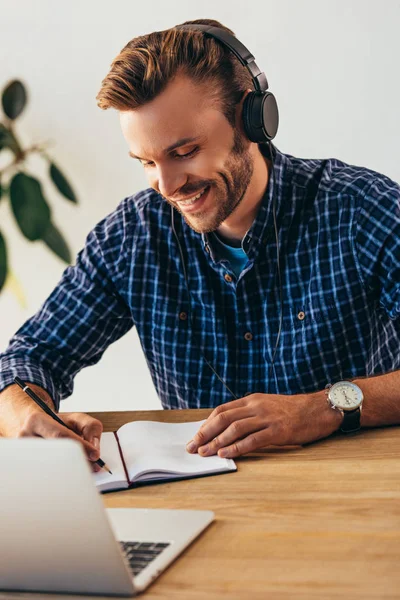 Retrato de homem sorridente em fones de ouvido fazendo anotações ao participar de webinar na mesa com laptop no escritório — Fotografia de Stock