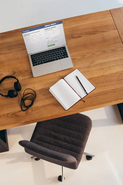 Top view of laptop with facebook website, headphones and notebook on wooden table with office chair near by — Stock Photo