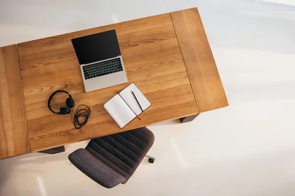 Вид зверху ноутбука з порожнім екраном, навушниками і блокнотом на дерев'яному столі з офісним кріслом поруч — стокове фото
