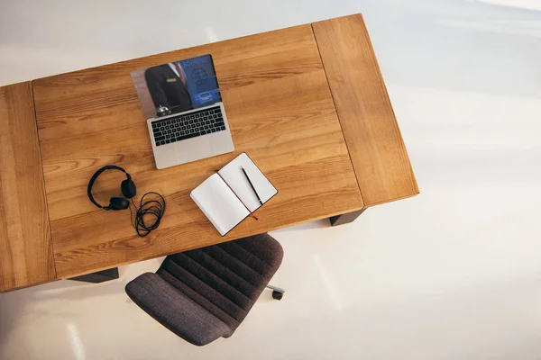 Top view of laptop with booking lettering on screen, headphones and notebook on wooden table with office chair near by — Stock Photo