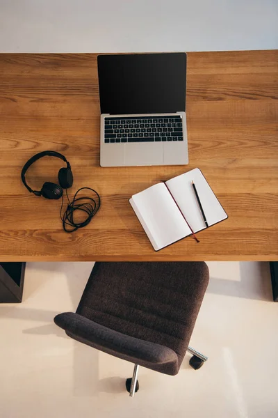 Vista superior do portátil com tela em branco, fones de ouvido e notebook na mesa de madeira com cadeira de escritório perto por — Fotografia de Stock