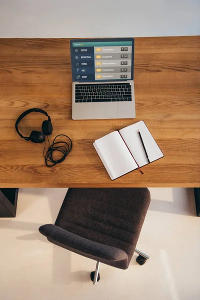 Вид зверху на ноутбук, навушники і блокнот на дерев'яному столі з офісним кріслом поруч — стокове фото
