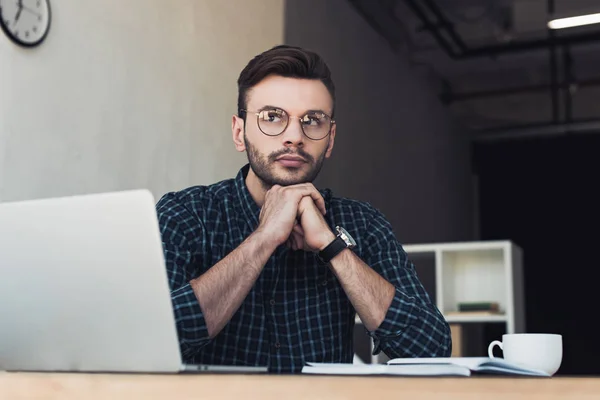 Portrait of pensive businessman in eyeglasses at workplace with laptop and notebook — Stock Photo