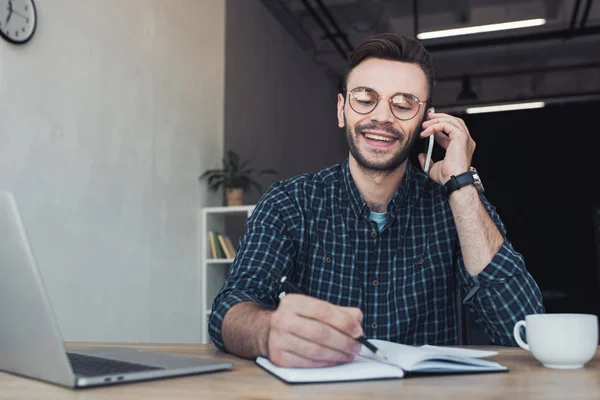 Портрет улыбающегося бизнесмена, разговаривающего на смартфоне на рабочем месте с ноутбуком и ноутбуком — стоковое фото