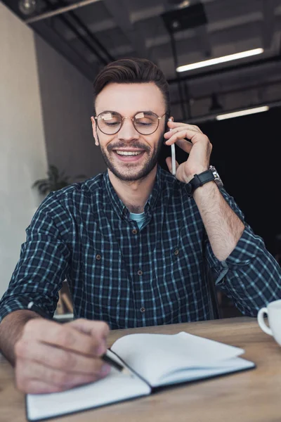 Porträt eines lächelnden Geschäftsmannes, der am Arbeitsplatz mit dem Notebook auf dem Smartphone spricht — Stockfoto