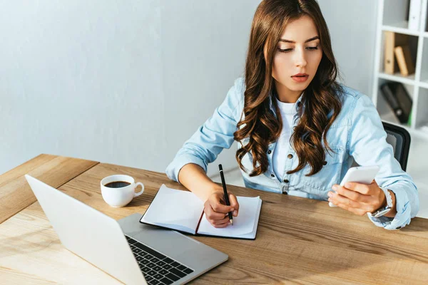 Junge Frau mit Smartphone am Tisch mit Laptop nimmt an Webinar im Büro teil — Stockfoto
