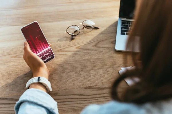 Partial view of businesswoman with smartphone with trading courses lettering on screen working on laptop at workplace — Stock Photo