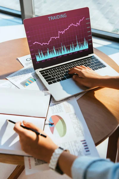 Partial view of businesswoman remote working on laptop with trading lettering on screen at workplace — Stock Photo