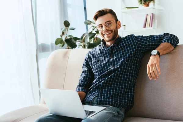 Retrato de homem sorridente em óculos com laptop sentado no sofá em casa — Fotografia de Stock
