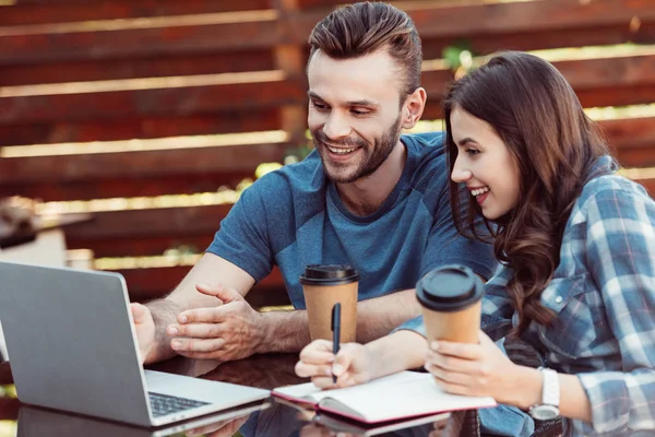 Cheerful friends at tabletop with laptop and notebook taking part in webinar together in cafe — Stock Photo