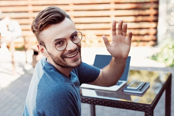 Sorridente bell'uomo che agita la mano e guarda la macchina fotografica al caffè di strada — Foto stock