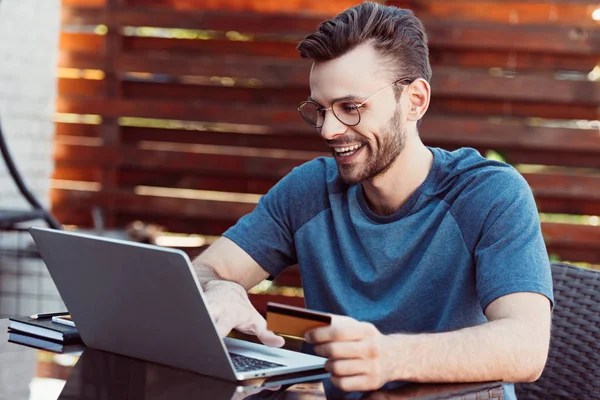 Sorrindo bonito homem compras on-line com cartão de crédito e laptop à mesa no café de rua — Fotografia de Stock