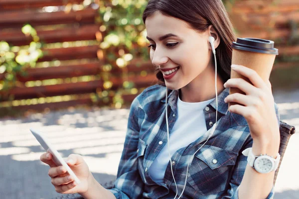 Lächelnde attraktive Frau, die mit Smartphone auf der Straße Musik hört und Kaffee in Pappbecher hält — Stockfoto